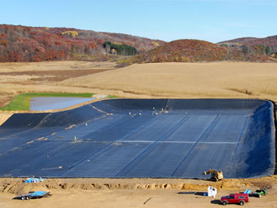 Some workers are laying the black geomembrane liners on a huge area, and there are some cars beside it.