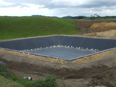 A huge square pit is wrapped with black PVC geomembrane, and there is grasslands beside it.