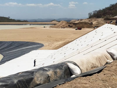 Rolls of white geosynthetic clay liner are laid on the slope, black geomembrane is covered on it, and there are some workers working.
