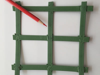 A piece of green biaxial geogrid is lying on the ground, and a red pencil is on it for contrast.