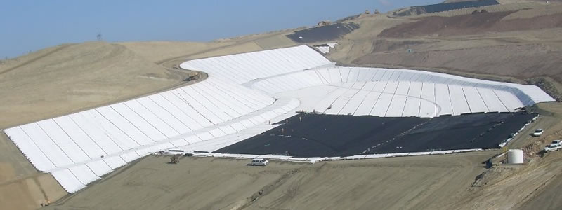 White geosynthetic clay liner and black geomembrane are laid on a large area, and there are some cars beside it.